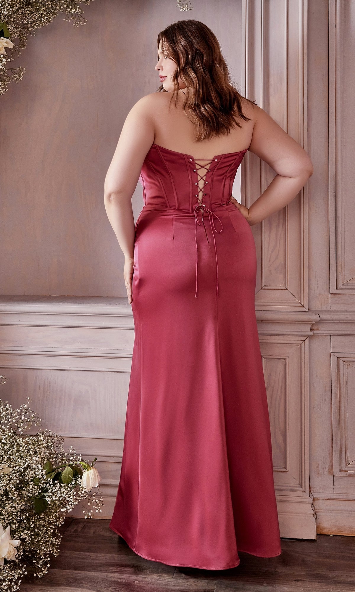 A woman stands indoors, facing away from the camera, wearing the Off-the-Shoulder Long Plus-Size Prom Dress 7484C in a stunning rose color with a lace-up back.